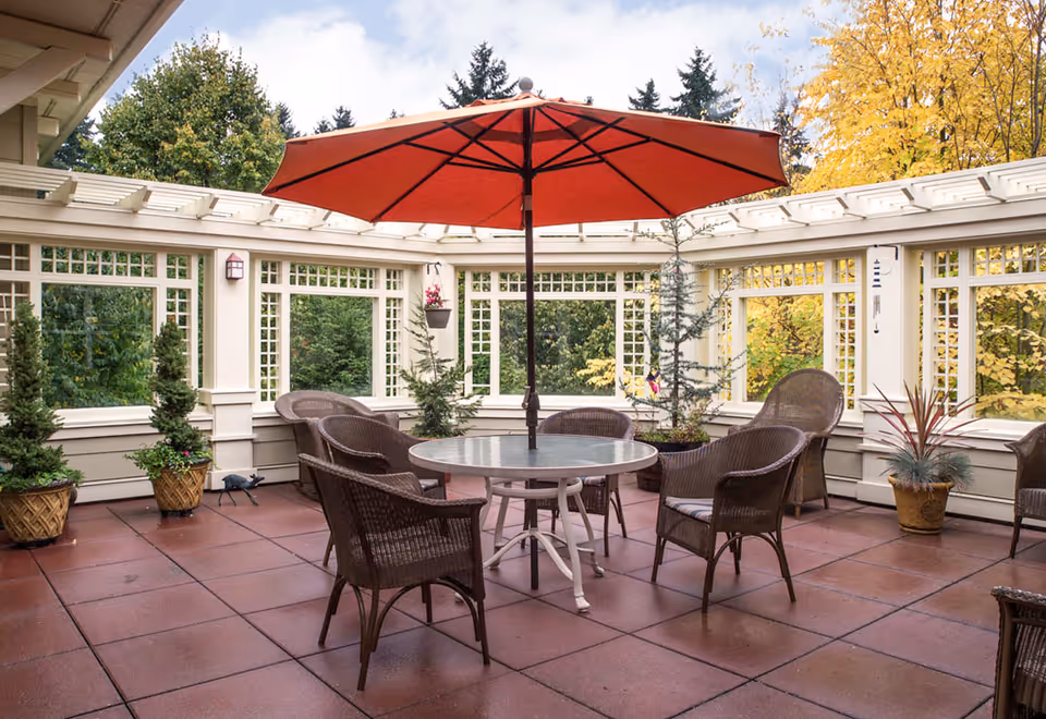 Outdoor patio area with a round glass table and six wicker chairs arranged around it. A large red umbrella is centered over the table. The patio is surrounded by white lattice fencing with windows, and there are several potted plants and small trees around the perimeter. Trees with green and yellow leaves are visible outside the patio.