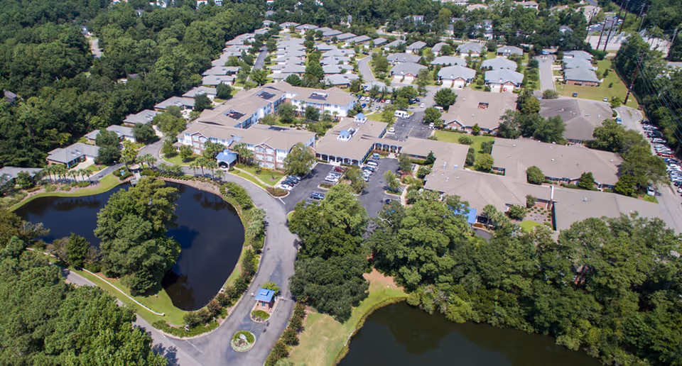 Aerial view of The Lakes at Litchfield senior living facility showing multiple buildings with beige roofs surrounded by trees, parking lots with cars, and two small lakes with greenery around them.