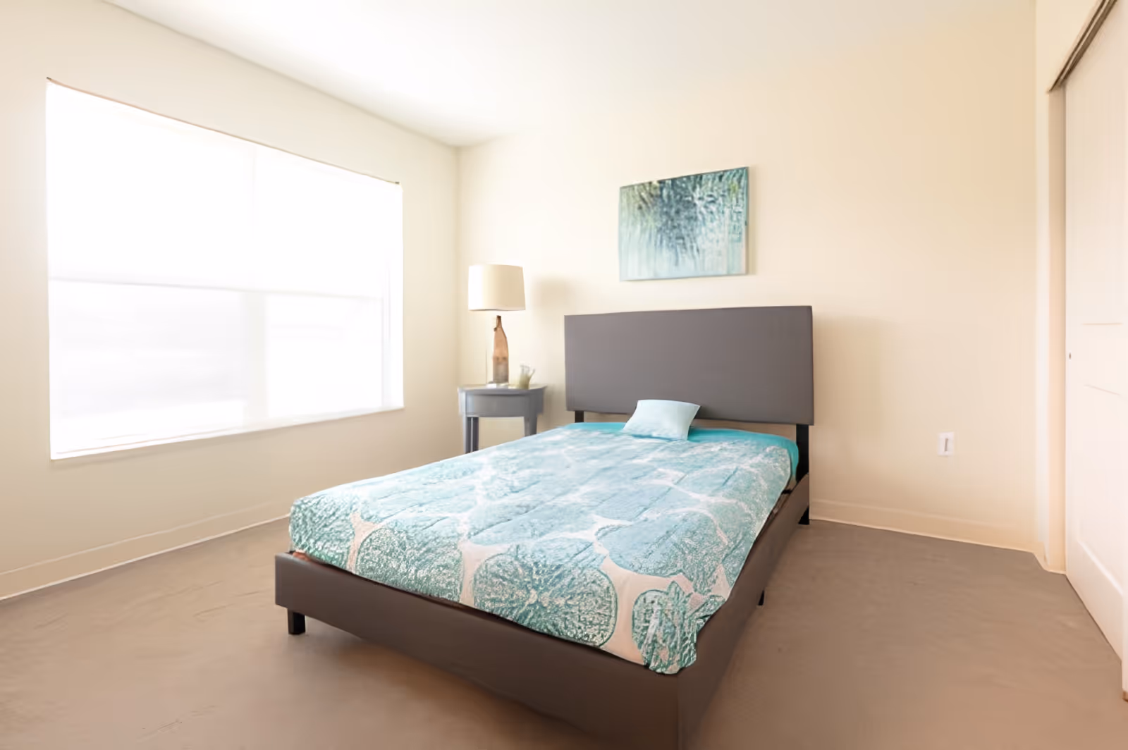 A bright bedroom with a large window covered by a white blind, a bed with a gray headboard and a teal patterned bedspread, a small round side table with a lamp and a decorative item, and a piece of abstract artwork hanging above the bed.