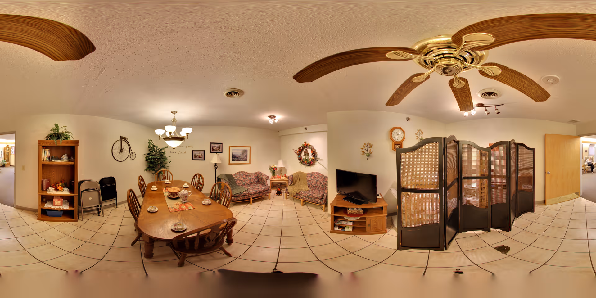 A combined dining and sitting room with a wooden dining table set, upholstered couches, a TV on a stand, decorative wall art, and ceiling fans.