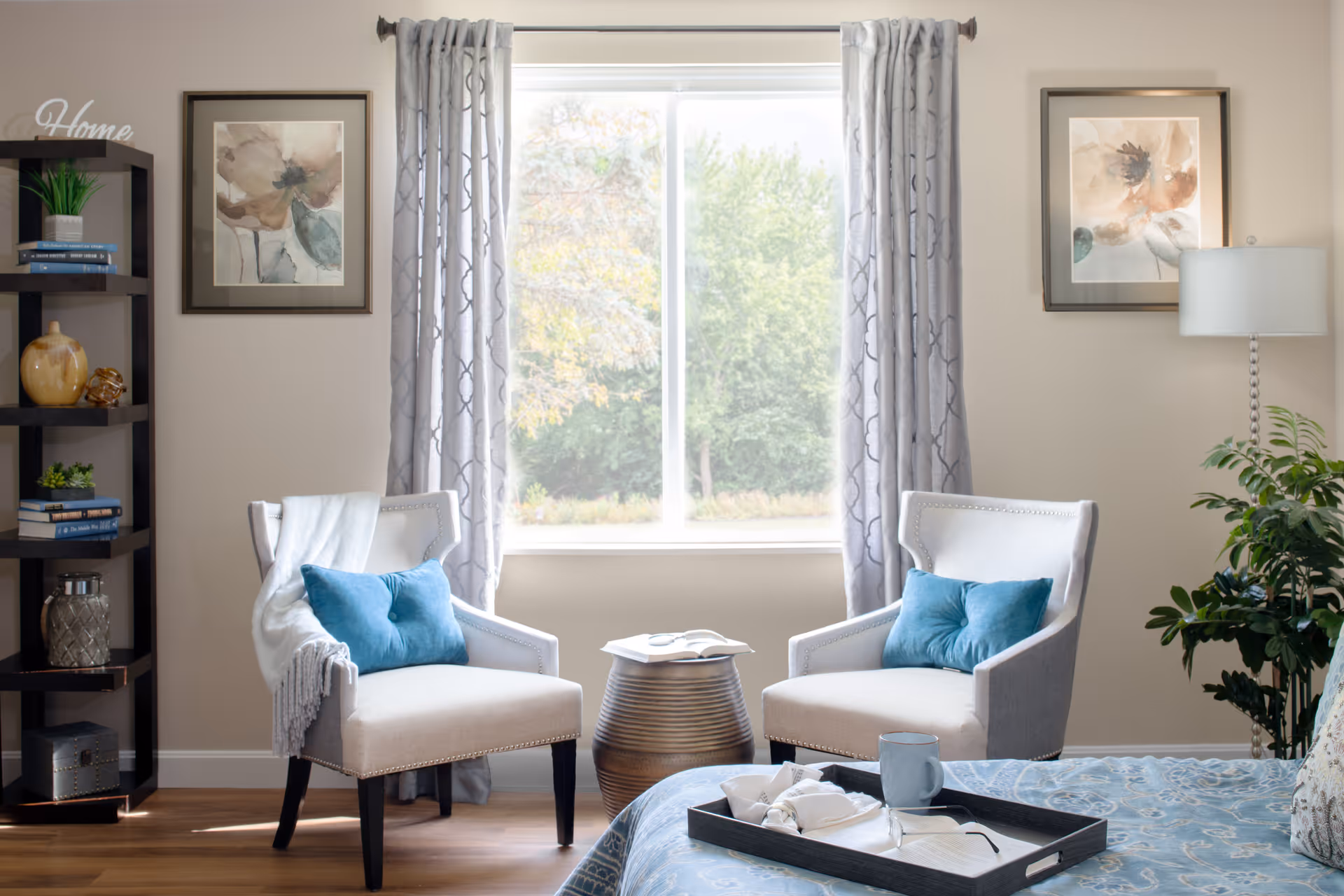 Sunlit bedroom seating area with two upholstered chairs by a window, shelving on the left and part of a bed with a tray in the foreground.