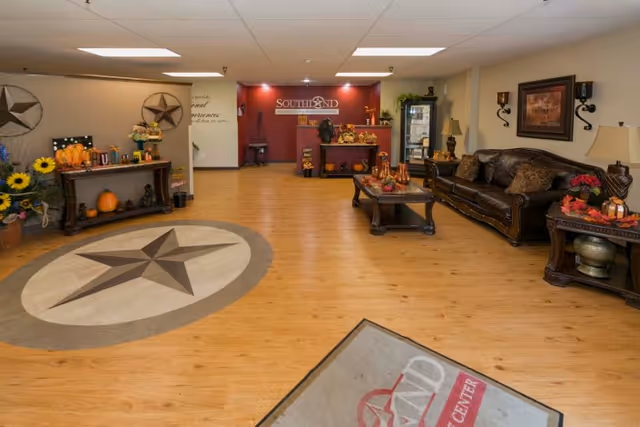 A spacious and warmly decorated lobby area with wooden flooring featuring a large star design. There is a dark brown leather sofa with cushions, a coffee table with decorative items, and side tables with lamps and fall-themed decorations. The back wall is painted red with the Southland Rehabilitation And Healthcare Center logo displayed. The area also includes a console table with pumpkins, flowers, and other seasonal decor.