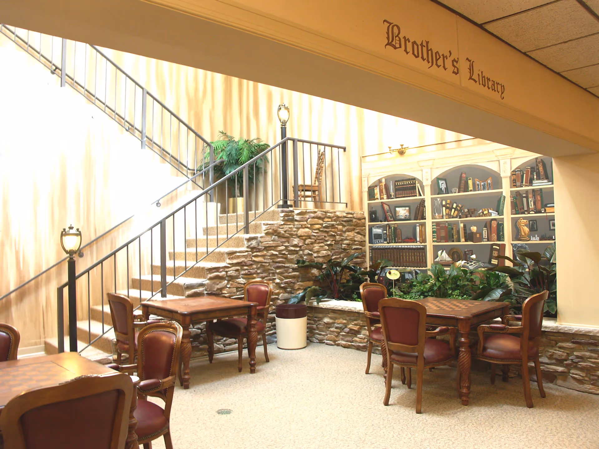 Cozy interior library/lounge with tables and chairs, a staircase, stone planter and bookshelves beneath a sign reading 'Brother's Library'.