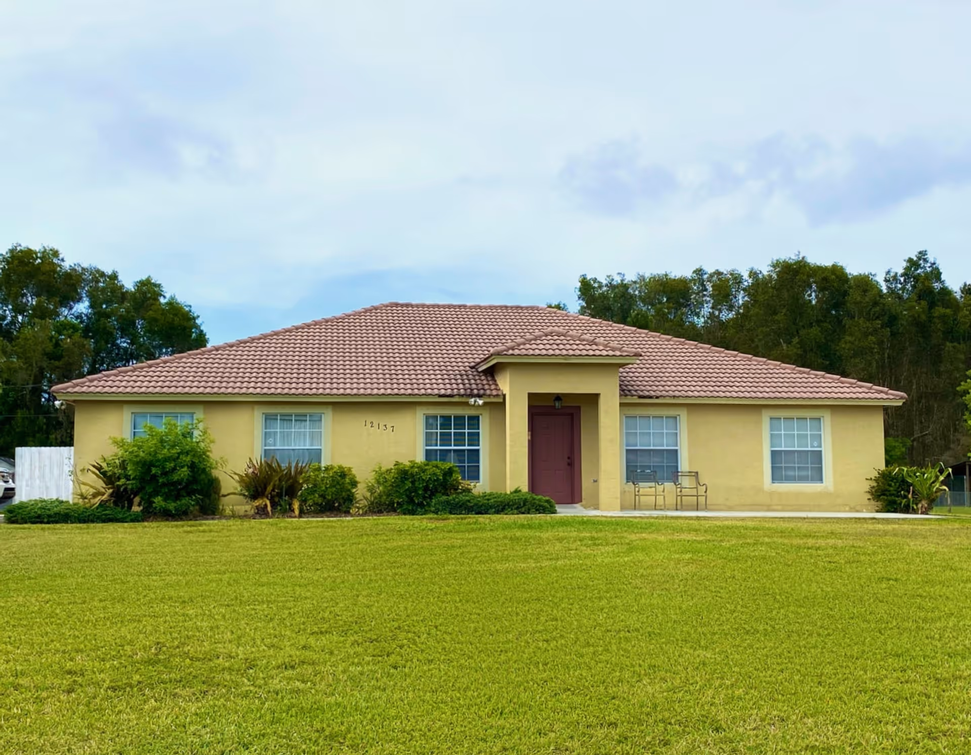 Front view of a single-story beige house with a tiled roof, red front door, windows, and a large lawn.