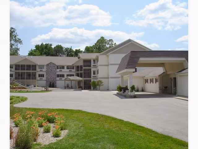 Exterior view of a senior living community building with a covered entrance, multiple windows, and a stone accent wall. The foreground features a paved driveway and a landscaped area with green grass and flowering plants under a partly cloudy sky.