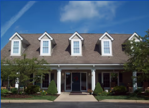 Front exterior view of a building with a sloped roof featuring five dormer windows, a covered porch with white columns, and a central entrance door. The landscaping includes small bushes, trees, and a well-maintained lawn under a blue sky with some clouds.