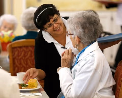 A smiling caregiver converses with an elderly woman at a dining table with a plate of food and a cup.