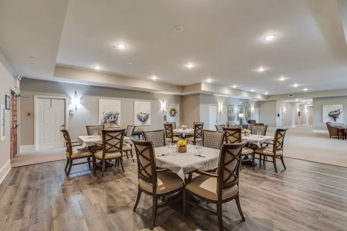 Spacious dining room in an assisted living facility with multiple set tables and chairs and soft overhead lighting.