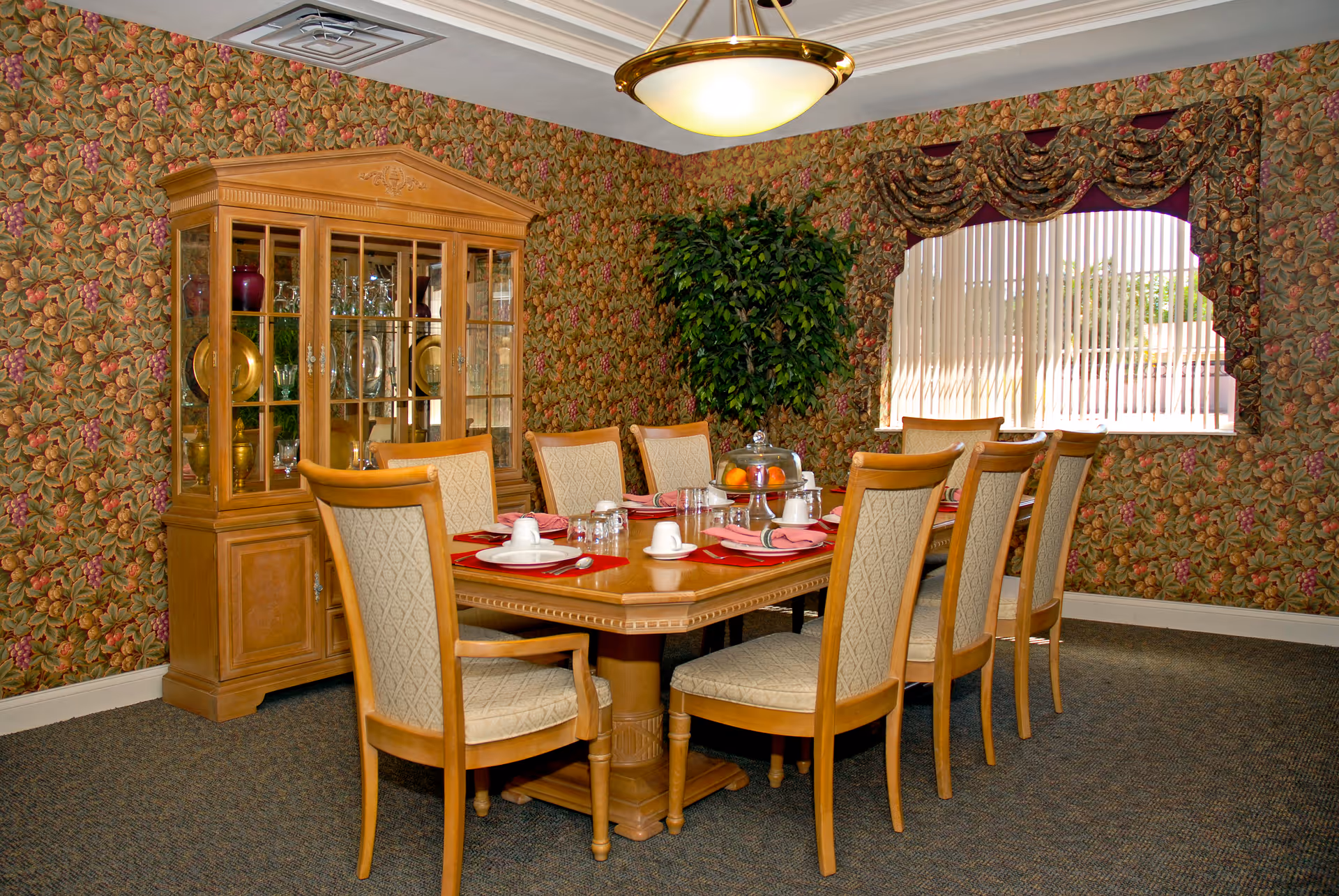 A dining room with a wooden dining table set for eight people. The table has white plates, cups, glasses, and red placemats. There is a wooden china cabinet with glass doors displaying dishes and glassware. The walls are covered with floral patterned wallpaper, and a large window with vertical blinds and decorative valances lets in natural light. A green potted plant is placed in the corner, and a ceiling light fixture hangs above the table.