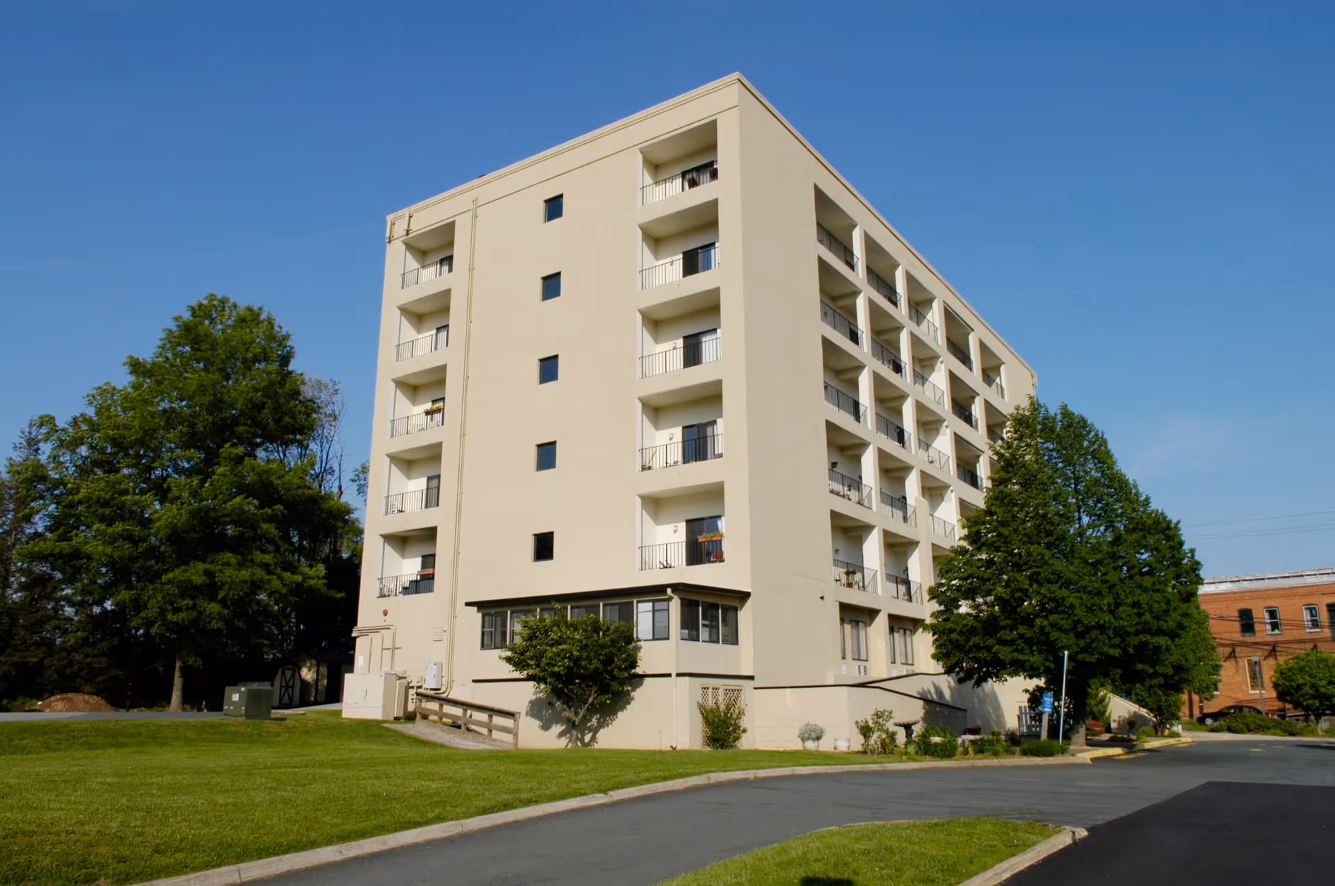 Exterior view of a multi-story beige building with balconies on each floor, surrounded by green trees and a well-maintained lawn under a clear blue sky.