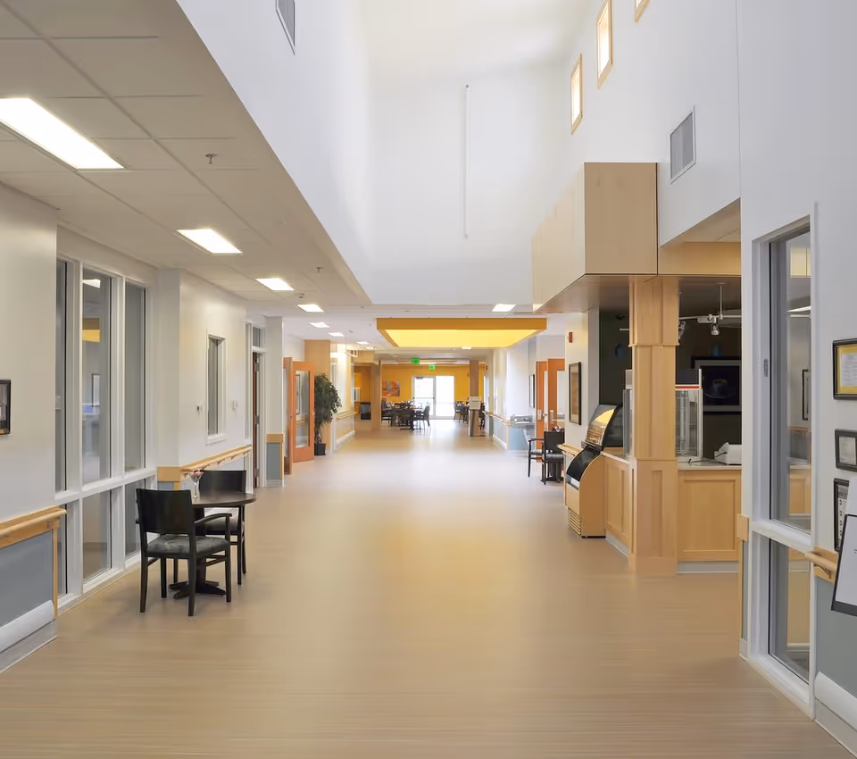 A wide, bright hallway in a senior living facility with light wood flooring and white walls. There are several doors and windows along the hallway, a small table with two chairs on the left side, and a wooden reception or service counter on the right. The ceiling has recessed lighting and a yellow accent panel near the far end. The hallway leads to a dining area with tables and chairs visible in the distance.