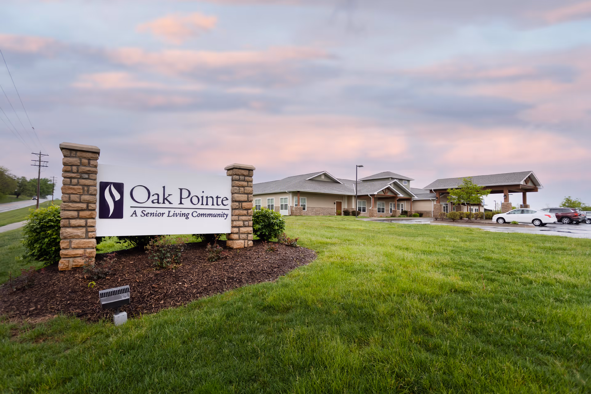 Exterior view of Oak Pointe senior living community building with a large sign in the foreground reading 'Oak Pointe A Senior Living Community'. The building is surrounded by green grass and a few parked cars under a cloudy sky at sunset.