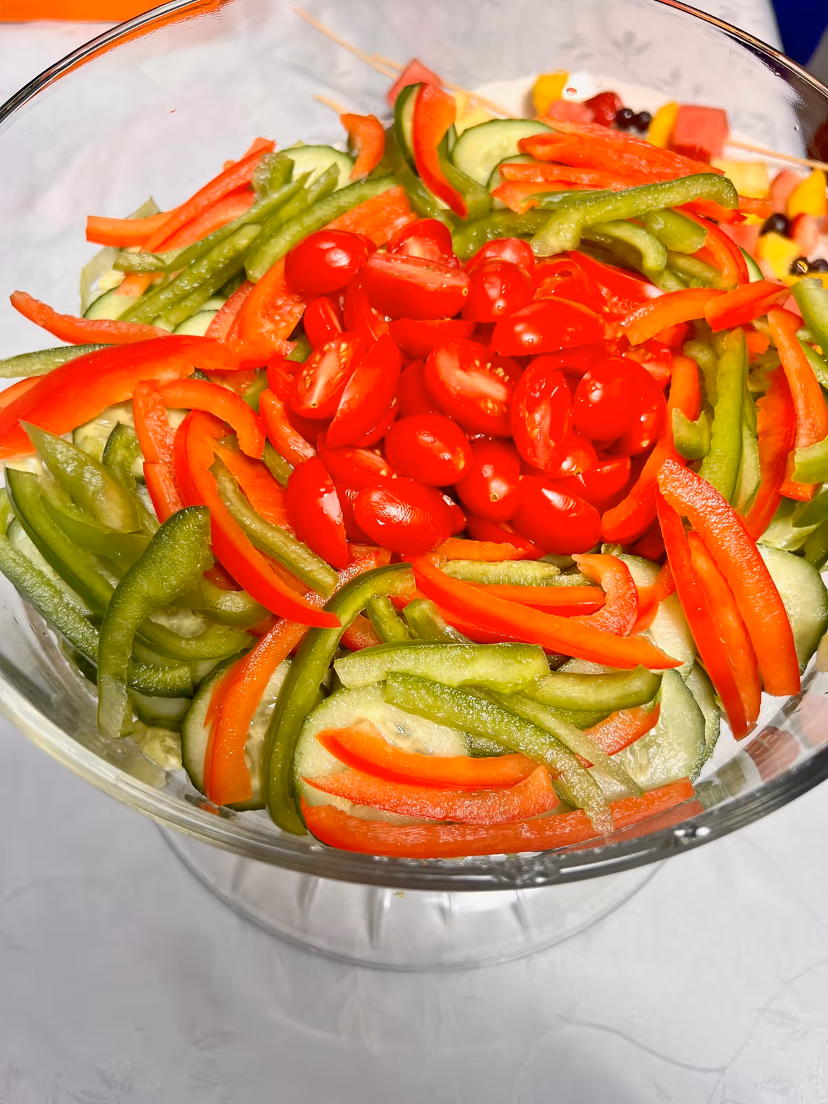 A clear glass bowl filled with a fresh vegetable salad containing sliced cucumbers, green and red bell peppers, and halved cherry tomatoes arranged on top.