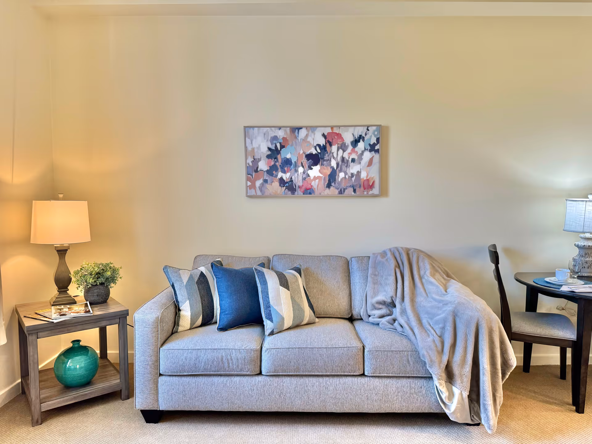 A cozy living room area with a light gray sofa adorned with three decorative pillows in shades of blue and gray, and a soft gray throw blanket draped over one side. Above the sofa hangs a colorful abstract floral painting. To the left of the sofa is a wooden side table with a lamp, a small green plant, and a decorative teal vase on the lower shelf. To the right is a small round table with a chair, a lamp, and a cup and saucer on the table. The walls are painted a light beige color and the floor is carpeted.