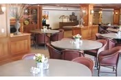 A dining room with round tables and maroon chairs arranged neatly. Each table has a small floral centerpiece. The room features wood paneling, large mirrors, and soft lighting, creating a warm and inviting atmosphere.