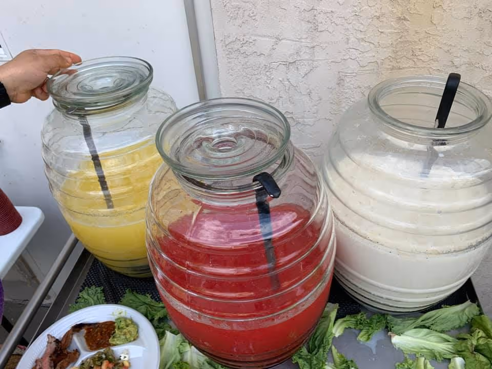 Three large glass beverage dispensers holding yellow, red, and white drinks on a serving table next to a plate of food.