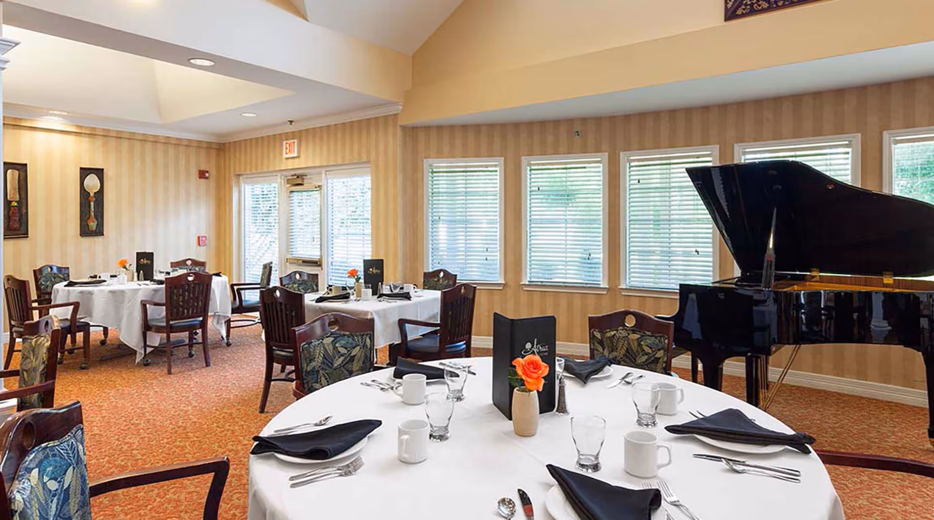 A dining room with round tables covered in white tablecloths, set with black napkins, white plates, cups, and silverware. There are wooden chairs with patterned cushions around the tables. A black grand piano is positioned near a wall with several windows covered by blinds. The walls have a beige striped wallpaper and framed artwork, and the floor is carpeted in a reddish pattern.