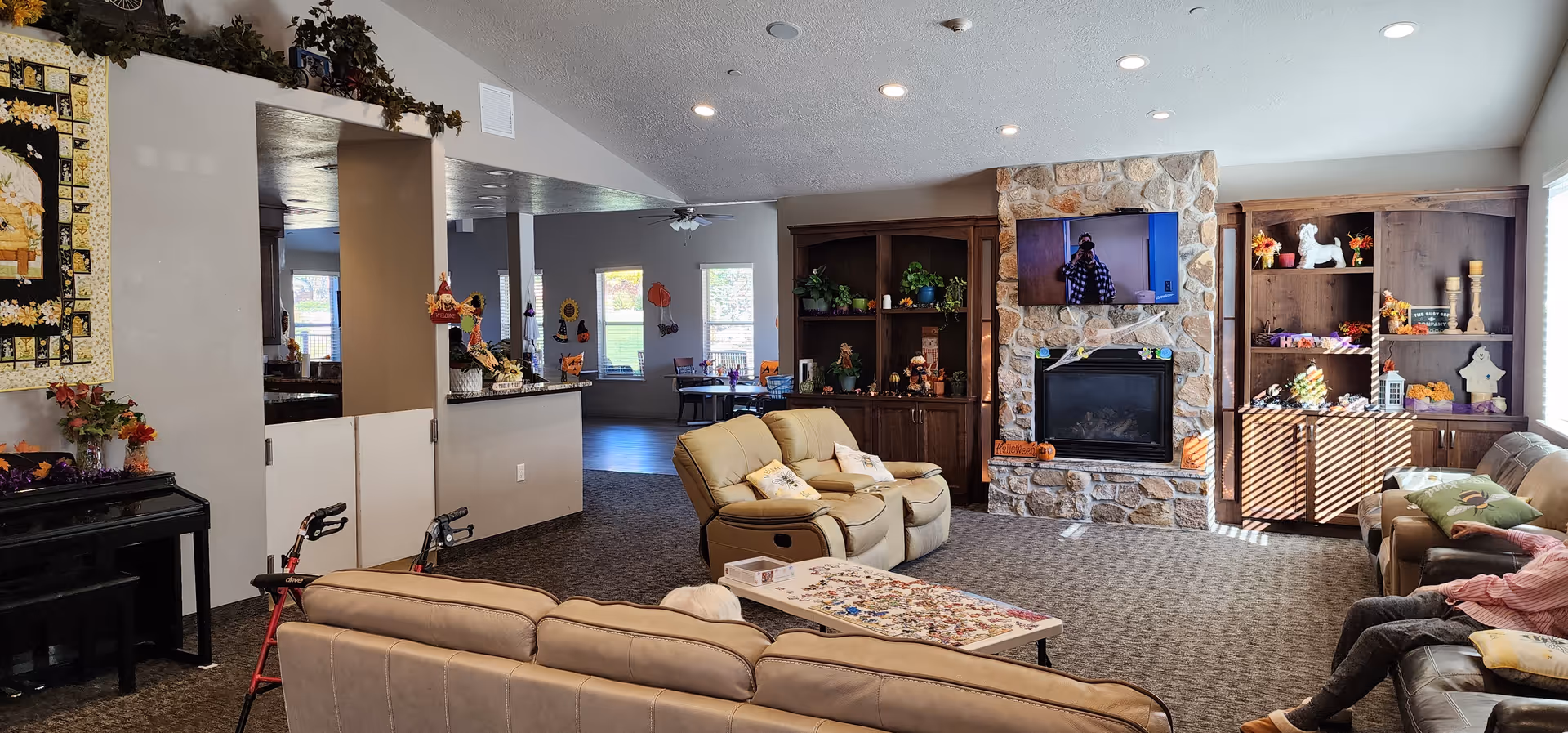 A spacious living room area in a senior living facility featuring beige leather sofas and recliners arranged around a coffee table with a puzzle on it. The room has a stone fireplace with a mounted flat-screen TV above it, flanked by wooden built-in shelves decorated with plants and seasonal decorations. Large windows allow natural light to fill the room, and there is a piano on the left side near a wall with a quilt hanging above it. The room is decorated with autumn-themed items, and a person is seated on the right sofa.