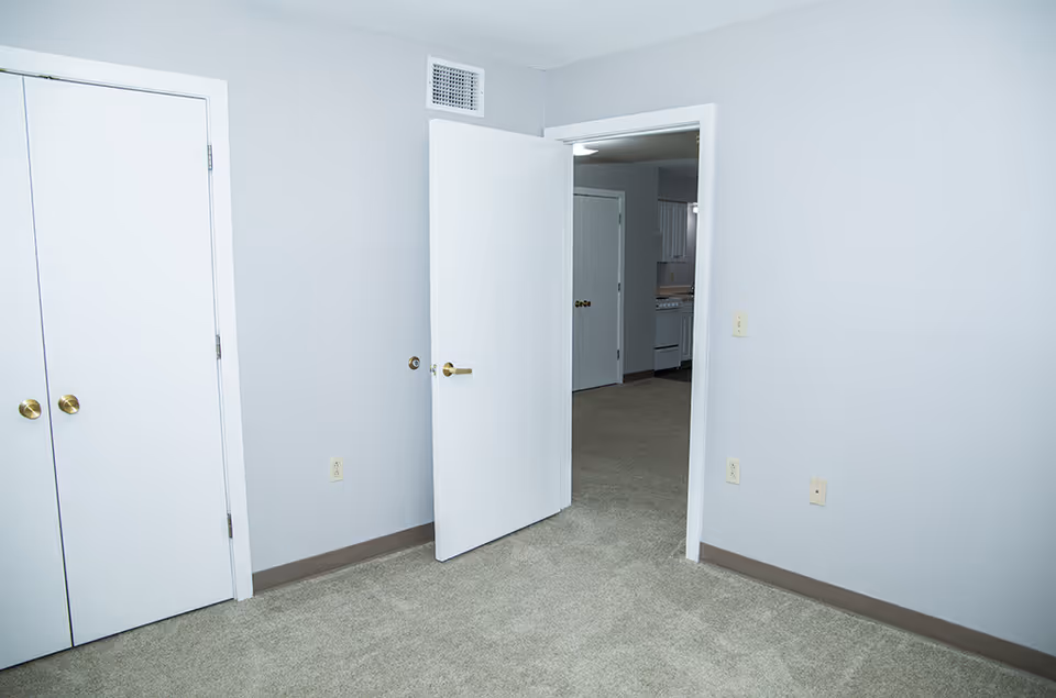 Empty carpeted room with a closed closet and an open door leading to a hallway and kitchen.