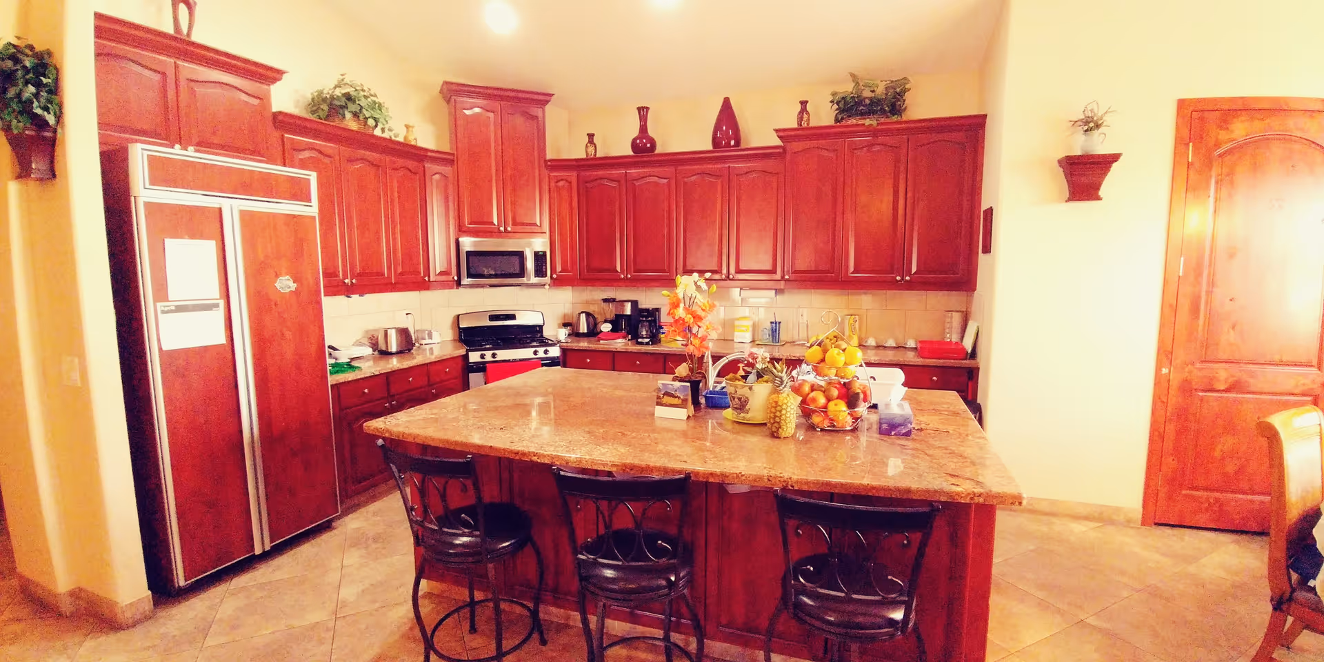 A spacious kitchen with dark wooden cabinets and a large central island topped with a marble countertop. The island has three black metal bar stools with cushioned seats. The kitchen features a stainless steel refrigerator, a microwave, a stove, and various small appliances on the countertops. There are decorative plants and vases on top of the cabinets and a bowl of fruit on the island. The floor is tiled, and there is a wooden door on the right side of the image.