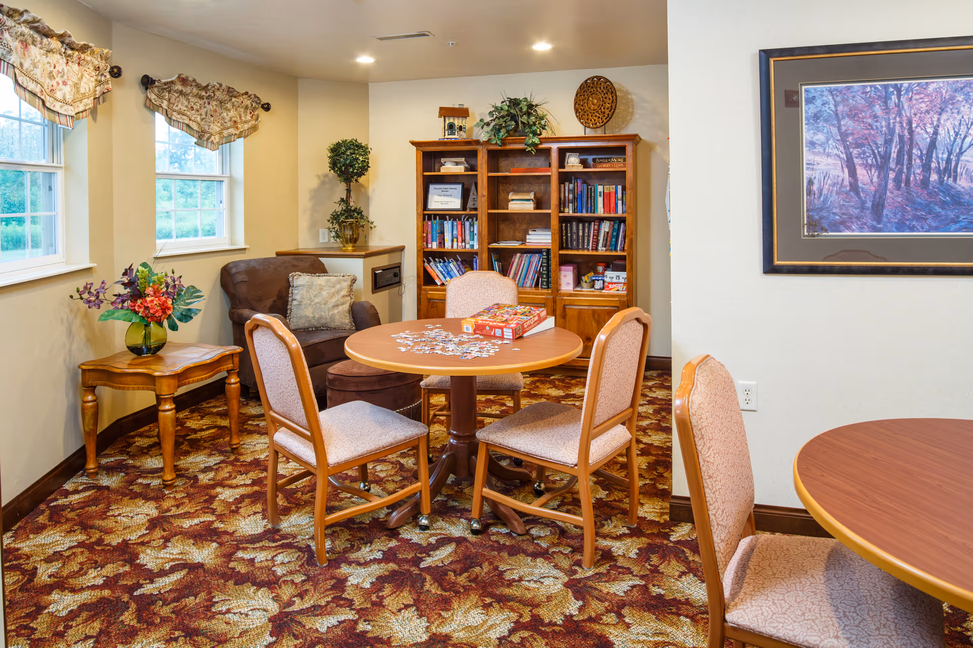 Cozy memory care common room with round tables and chairs, a jigsaw puzzle on the table, a bookshelf and an armchair by the windows.