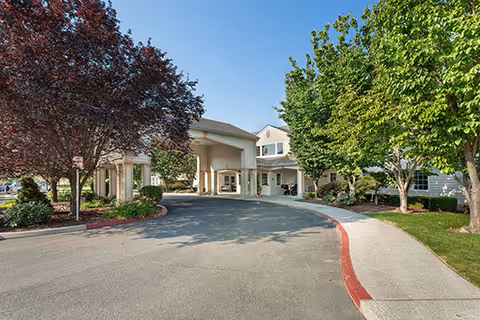 Exterior view of a senior living facility entrance with a covered drop-off area, surrounded by trees and landscaping under a clear blue sky.