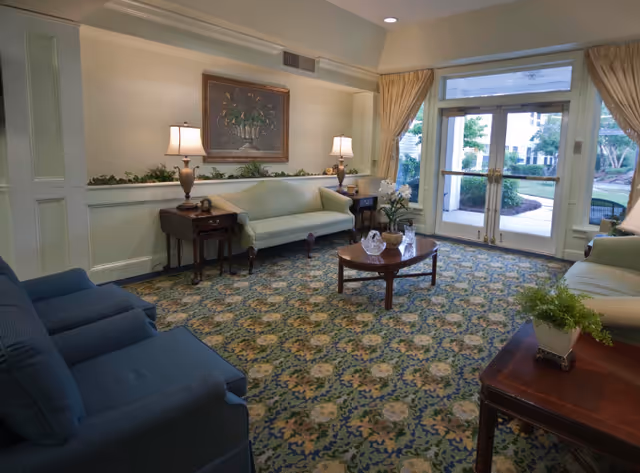 A cozy senior living facility common area with patterned carpet, two blue armchairs, two light green sofas, a wooden coffee table with decorative items, side tables with lamps, a framed floral painting on the wall, and large glass doors leading outside.