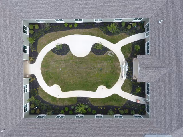 Aerial view of a rectangular courtyard enclosed by a building with a shingled roof. The courtyard features a curved concrete walkway surrounding a grassy area with small plants and shrubs along the edges. There is a small covered structure on one side of the courtyard.