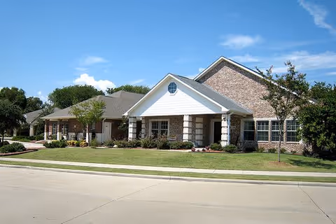 Exterior view of a single-story senior living facility building with brick and siding facade, a well-maintained lawn, small trees, and a clear blue sky.