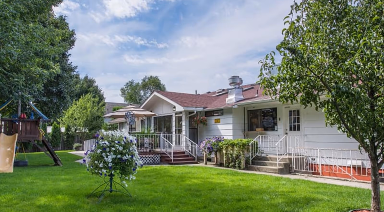 Single-story white care building with porches and ramps set behind a green lawn, flowering planters, and a children's playset under a partly cloudy sky.
