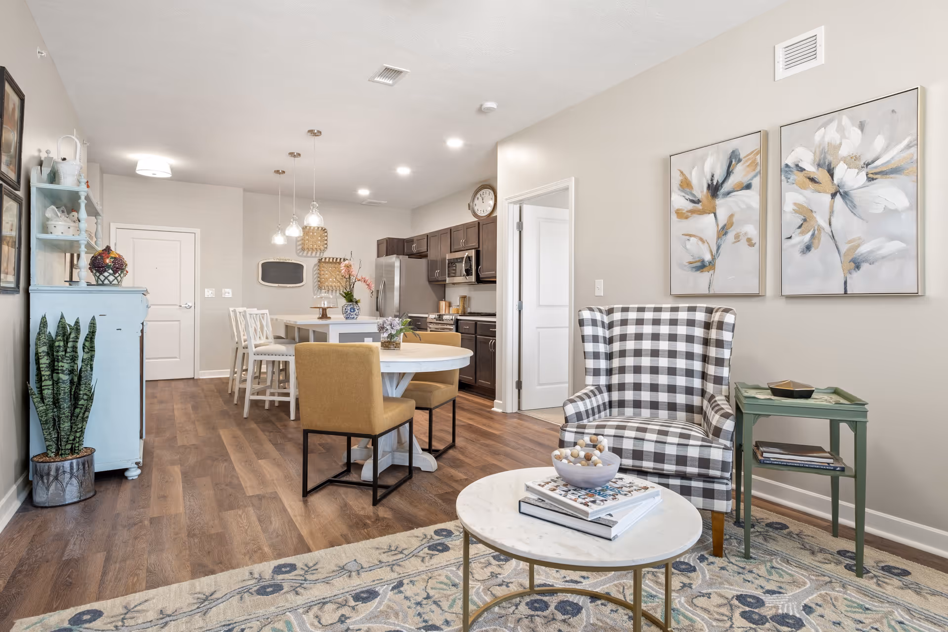 A bright and cozy senior living interior featuring a living area with a checkered armchair, a round marble coffee table with books and decorative items, and a green side table. The background shows a dining area with a round table and mustard yellow chairs, a kitchen with dark cabinets and stainless steel appliances, and light-colored walls adorned with floral paintings and wall decor.