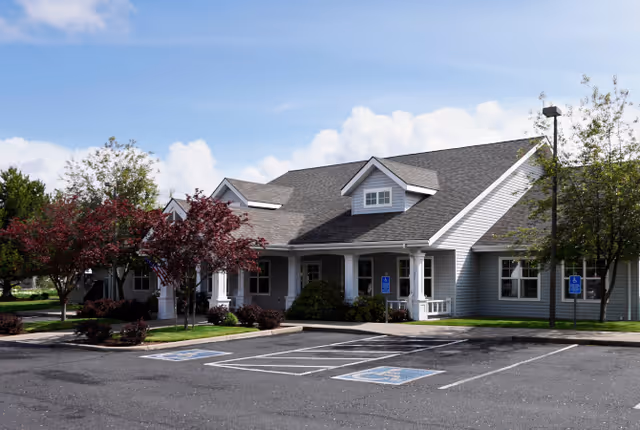 Single-story light-gray senior living facility with a covered front entrance, landscaped shrubs, and empty accessible parking spaces.