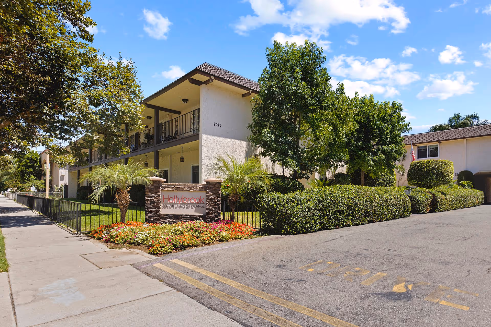 Front exterior of a two-story assisted living building with landscaped flower beds, palm trees, and an entrance sign under a blue sky.