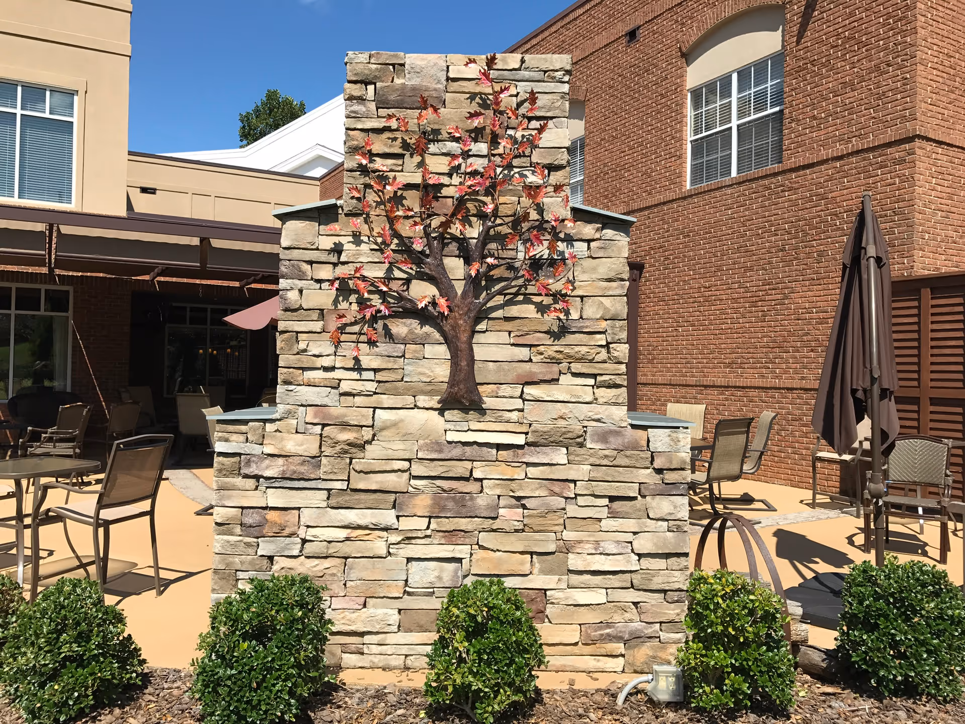 Outdoor patio area at Trinity Oaks featuring a stone wall with a metal tree sculpture mounted on it. Surrounding the wall are green shrubs, patio tables and chairs, and closed umbrellas. The background includes brick and beige building walls under a clear blue sky.