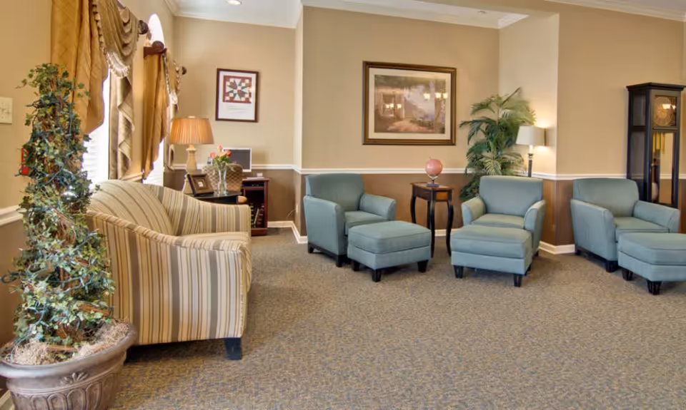 A cozy living room area with three light blue armchairs, one with an ottoman, and a striped loveseat. The room features beige walls with white trim, a carpeted floor, framed artwork on the walls, a tall plant, a small side table with a globe, a lamp, and a grandfather clock in the corner. There is also a potted plant with string lights near the window with draped curtains.