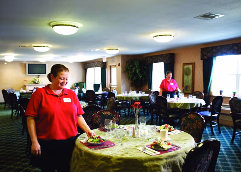 Two staff members in red shirts set and arrange place settings on round tables in a spacious senior living dining room.