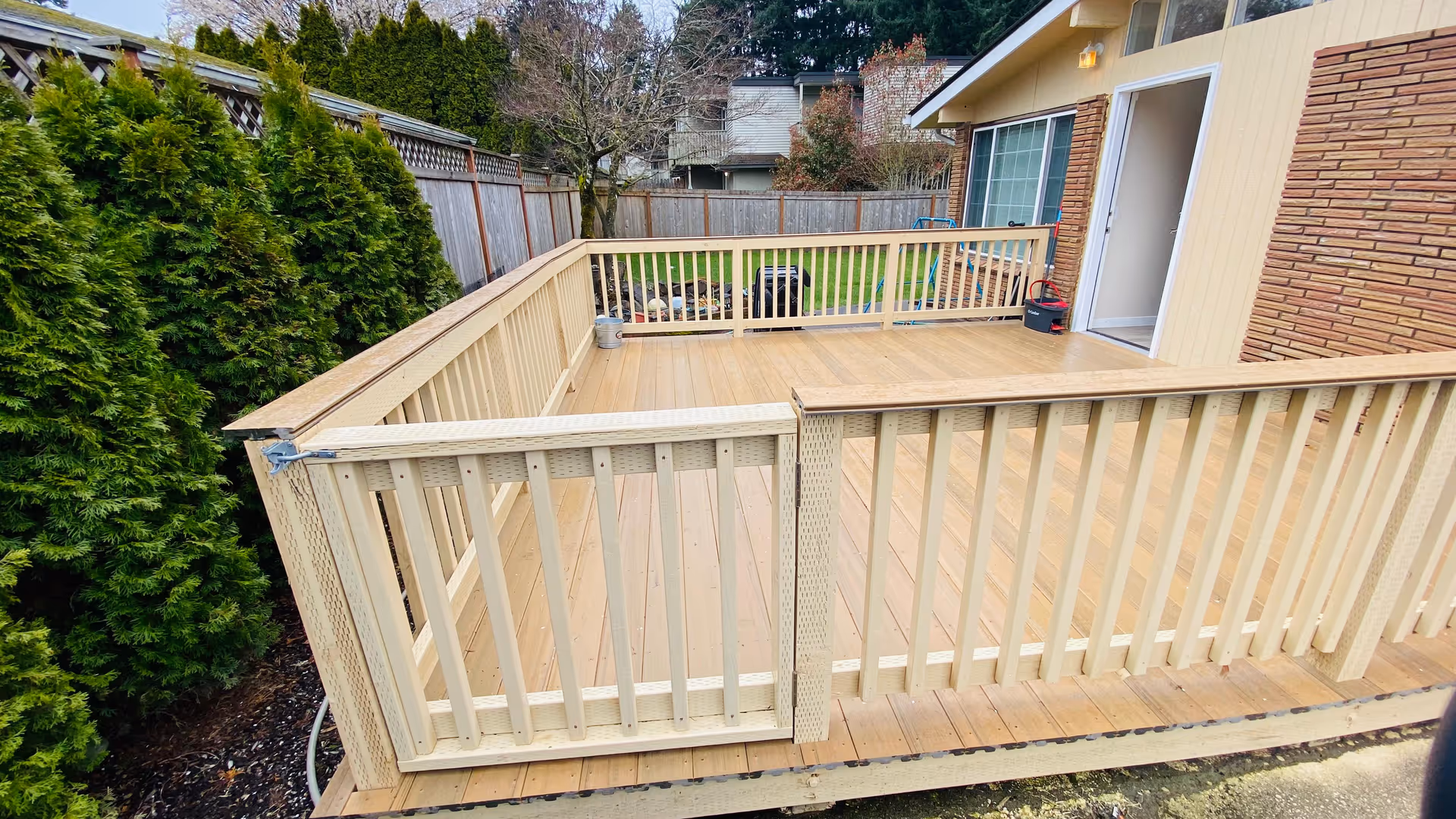 A wooden fenced deck attached to a house with beige siding and brick exterior. The deck has a gate and overlooks a backyard with green grass, trees, and a wooden fence. There are some outdoor items near the house wall.