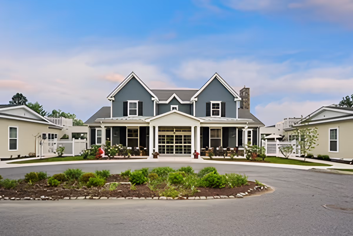 Front exterior view of a large two-story residential building with a gray facade, white trim, and black shutters. The building has a covered porch with several chairs and is flanked by two smaller single-story buildings. There is a circular driveway with a landscaped island in the foreground under a partly cloudy blue sky.