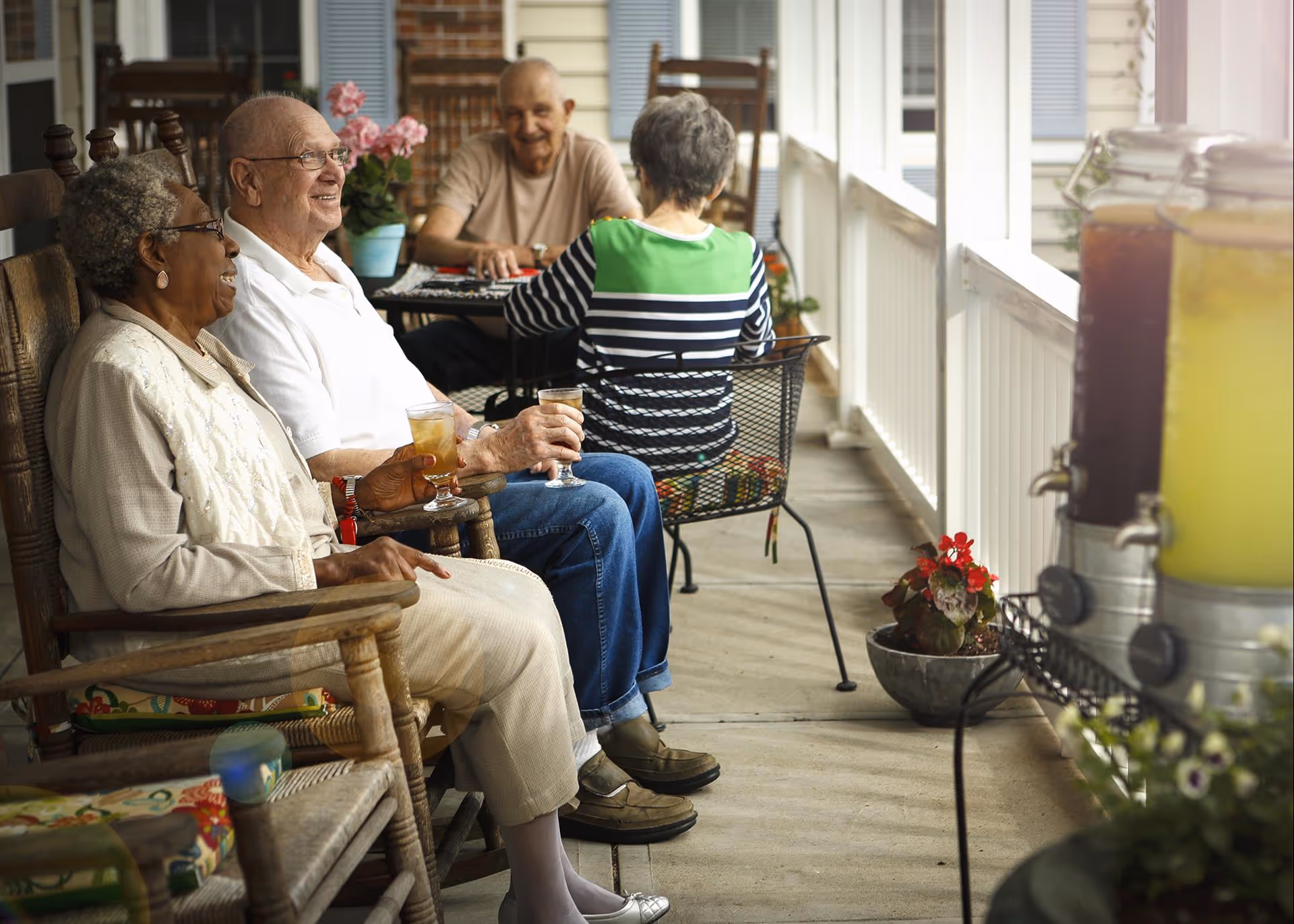Four elderly people sitting and enjoying drinks on a covered outdoor porch. Two are seated in wooden rocking chairs in the foreground, smiling and holding glasses of iced tea. Two others are seated at a small table in the background, engaged in conversation. The porch has potted plants and beverage dispensers with lemonade and other drinks on a stand to the right.