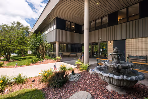 Exterior view of Oakwood Heights Village - Nursing and Rehabilitation Center showing the entrance area with a covered walkway supported by columns, a multi-tiered stone fountain surrounded by landscaping with small American flags, bushes, and trees under a partly cloudy sky.