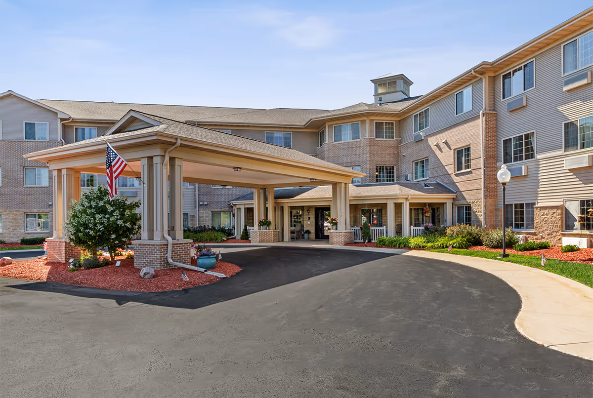 Exterior view of a senior living facility named LakeHouse Manitowoc featuring a covered entrance with columns, an American flag, landscaped flower beds with mulch, and a multi-story building with numerous windows under a clear blue sky.