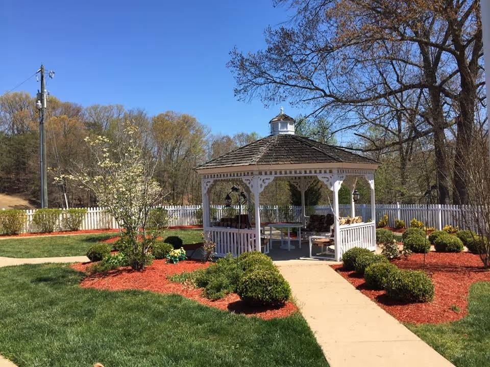 A white wooden gazebo with a shingled roof sits in a landscaped garden area with green grass, trimmed bushes, and red mulch. A concrete pathway leads to the gazebo, which contains outdoor seating and a table. Trees and a white picket fence surround the area under a clear blue sky.