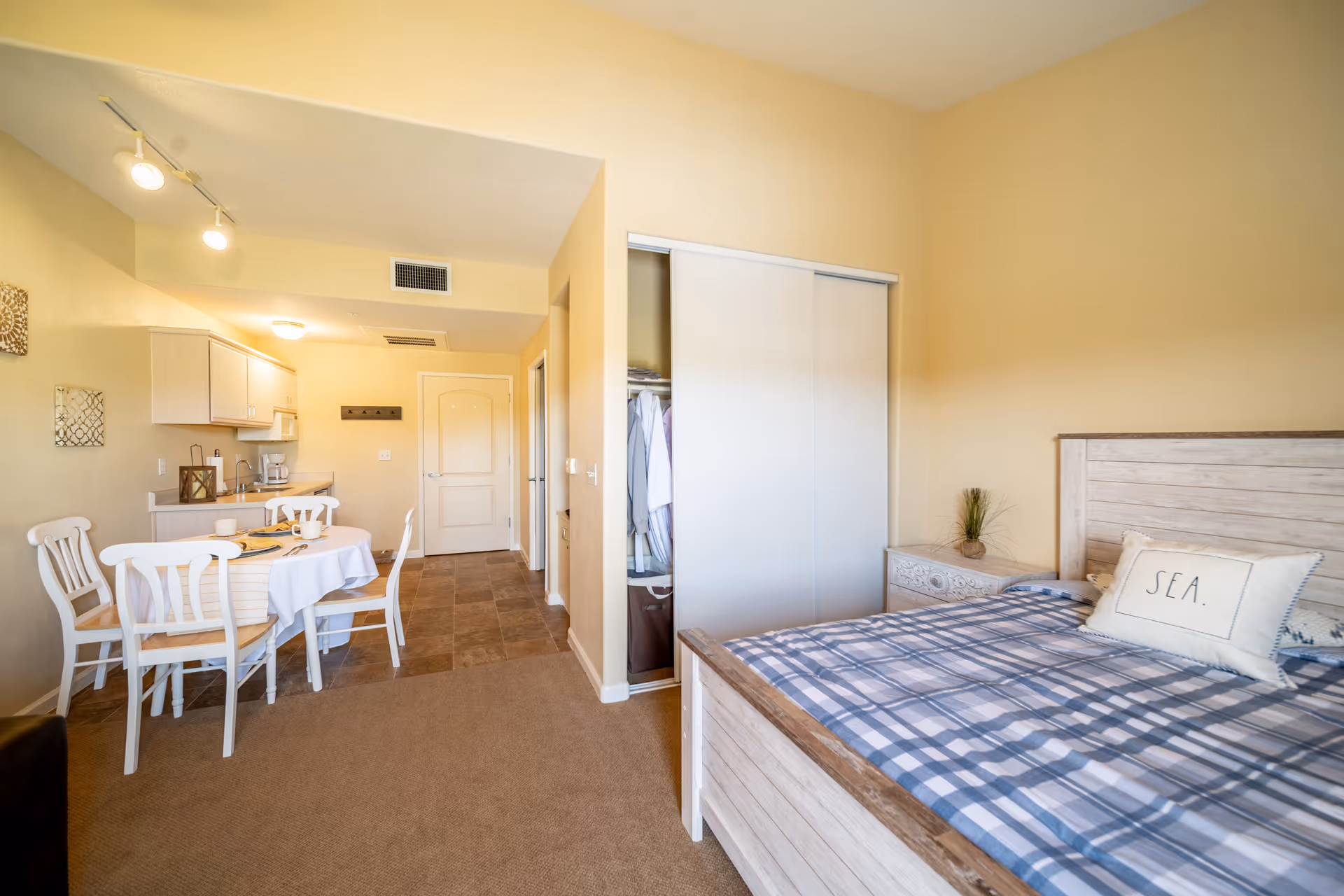 A cozy studio apartment interior featuring a bed with a blue plaid comforter and a pillow labeled 'SEA', a small nightstand with a plant, an open closet with clothes, a dining table set with four white chairs and tableware, and a compact kitchen area with cabinets, a sink, and a coffee maker. The walls are painted beige and the floor transitions from carpet in the sleeping area to tile in the kitchen and entryway.