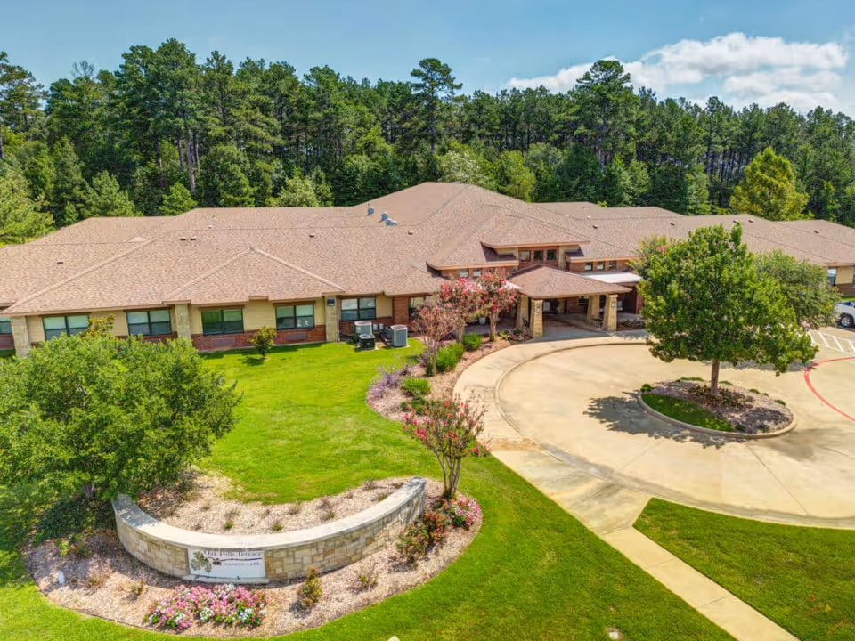 Aerial view of Oak Hills Terrace Memory Care facility showing a single-story building with a brown roof surrounded by green lawns, trees, and landscaped flower beds. There is a circular driveway with a tree in the center and a stone sign in front of the building.