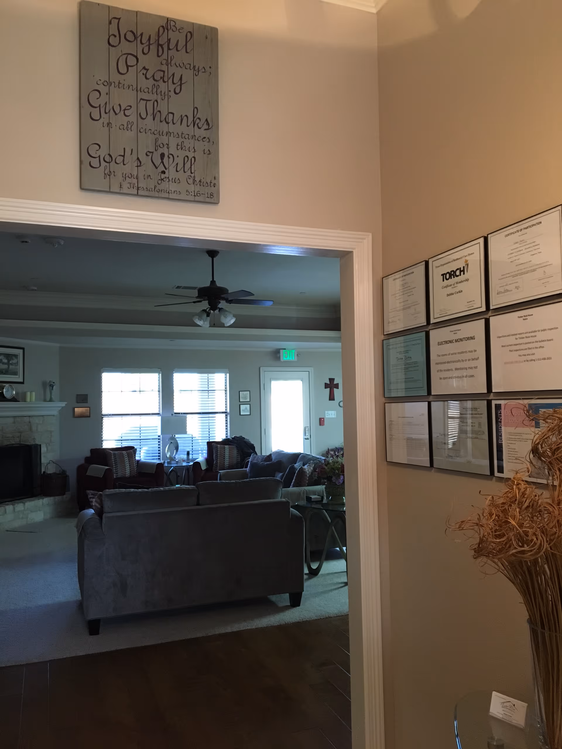 View through a doorway into a living room area with a gray couch, armchairs, a ceiling fan, and a fireplace. On the wall above the doorway is a wooden sign with an inspirational quote. To the right of the doorway, several framed certificates and notices are displayed on the wall, and a vase with dried decorative plants is visible.