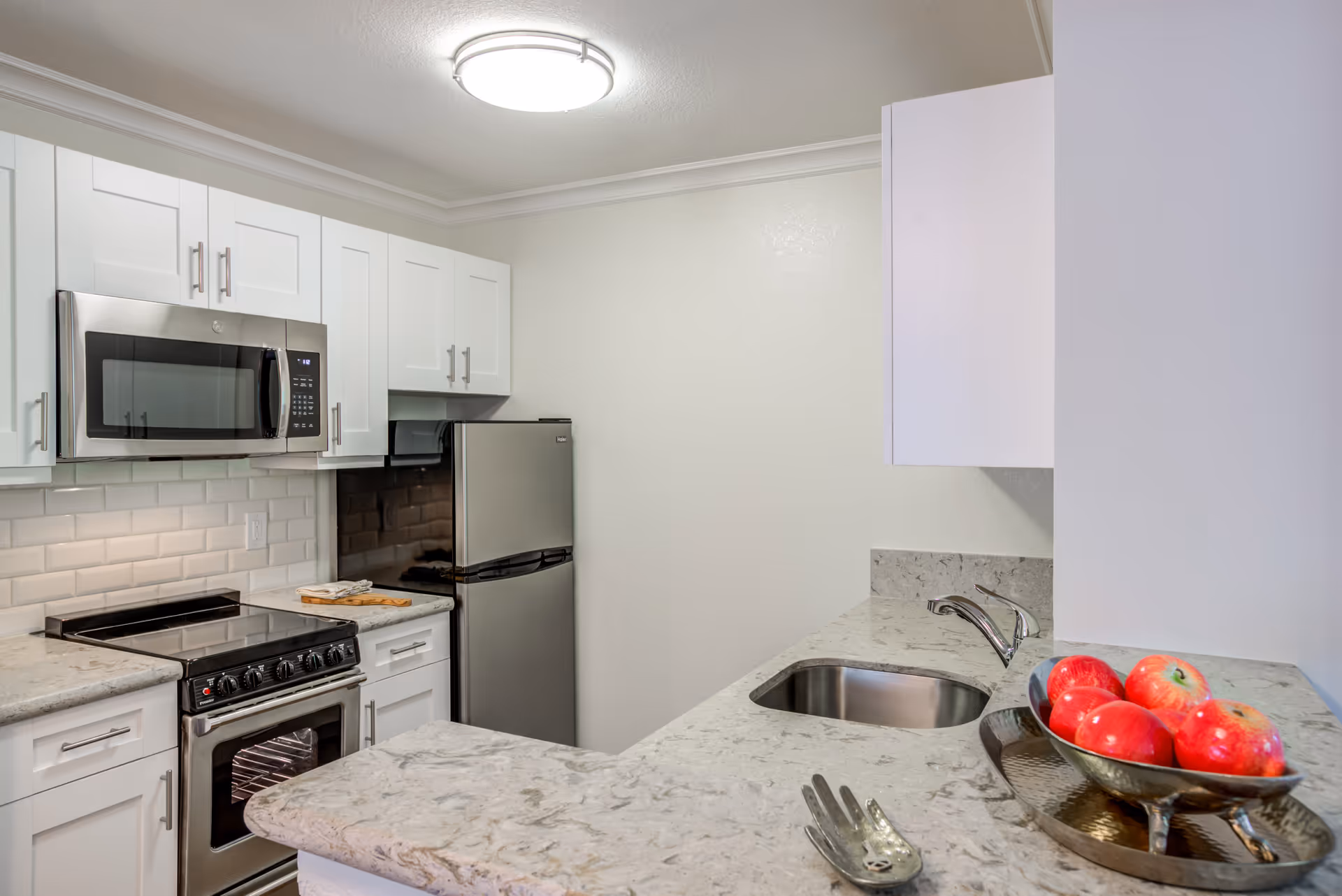 Modern kitchen with white cabinets, stainless steel microwave, stove, and refrigerator. Marble countertops with a sink and a bowl of red apples on the counter.