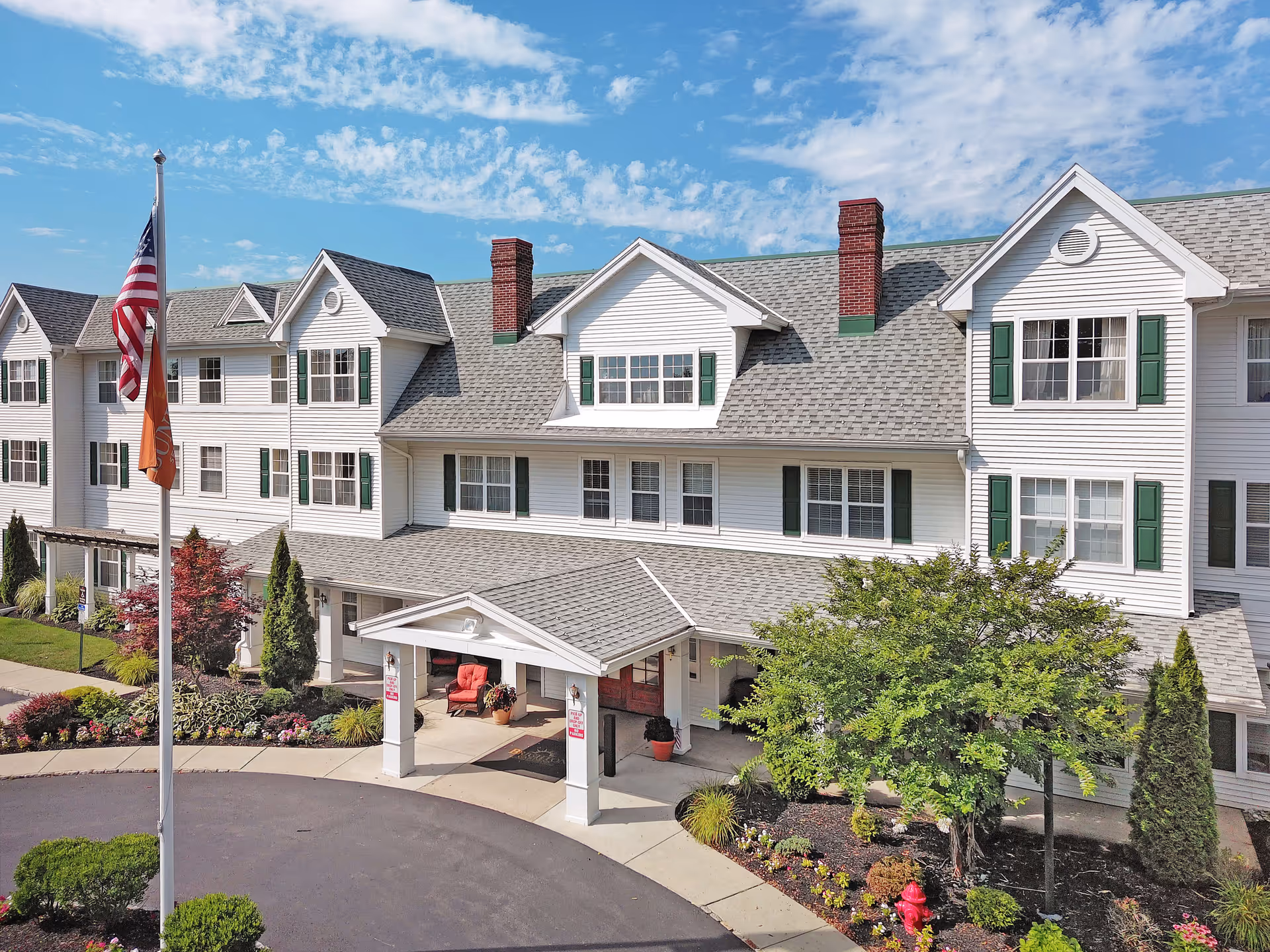Exterior front view of a large, white senior living facility building with green shutters and a gray shingled roof under a partly cloudy blue sky. The entrance has a covered porch with seating and is surrounded by landscaped bushes, trees, and flowers. Two flagpoles with flags are visible near the driveway.