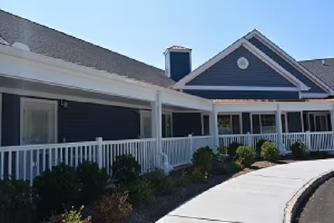 Blue-gray assisted living building with white porch railing and manicured shrubs along a curved path.