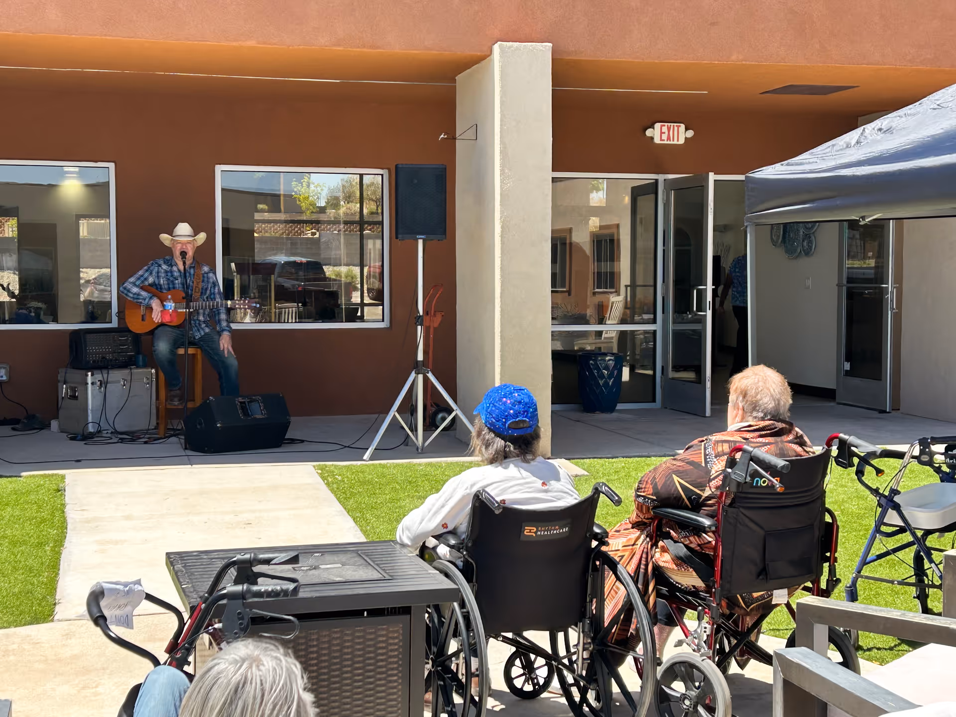 An outdoor courtyard area at Fountain Hill Assisted Living where a man wearing a cowboy hat and plaid shirt is playing guitar and singing into a microphone. Several elderly people in wheelchairs are seated on the grass facing the performer. The building has large windows and open double doors leading inside.