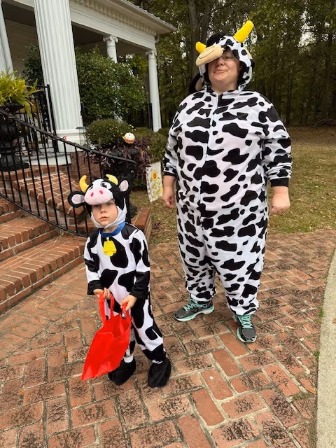 An adult and a child dressed in matching cow costumes standing outside on a brick pathway near a house with white columns and greenery in the background. The child is holding a red bag.